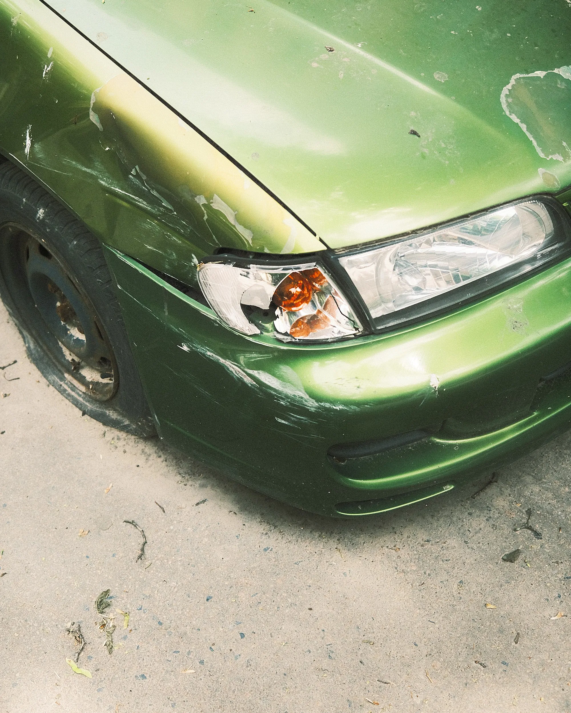 Front corner of a green car with a scuffed bumper and headlight